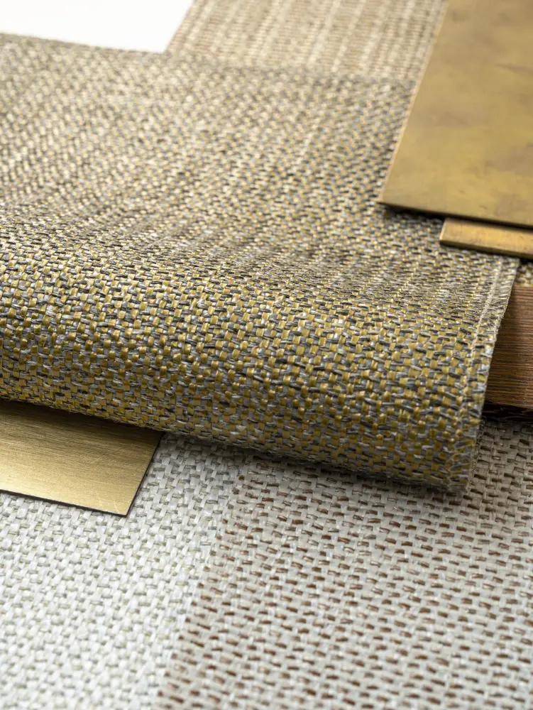 Close-up of Vescom Amber woven textile wallcovering samples with pronounced weave and irregular melange beside brass sheets