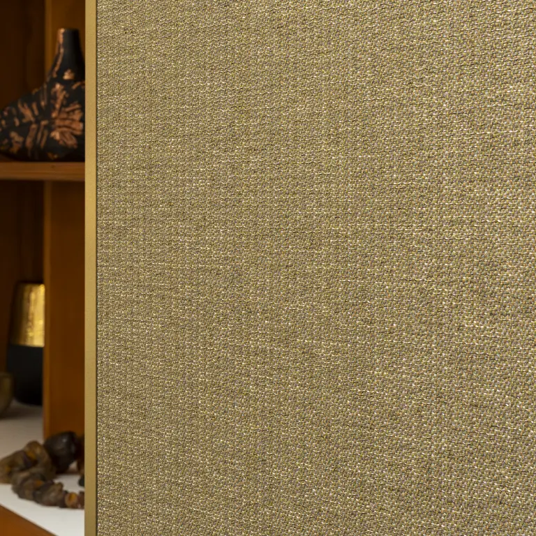 Vescom Amber woven textile wallcovering with warm gold tone and irregular melange texture beside a wooden shelving unit.