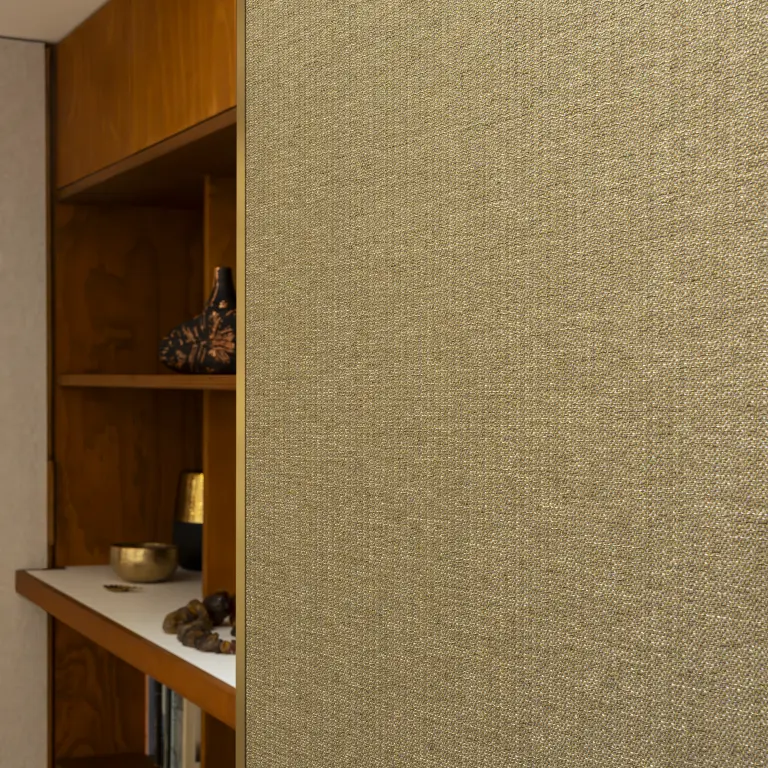 Close-up of Vescom Amber woven textile wallcovering with warm gold tone and irregular melange texture beside wooden shelving