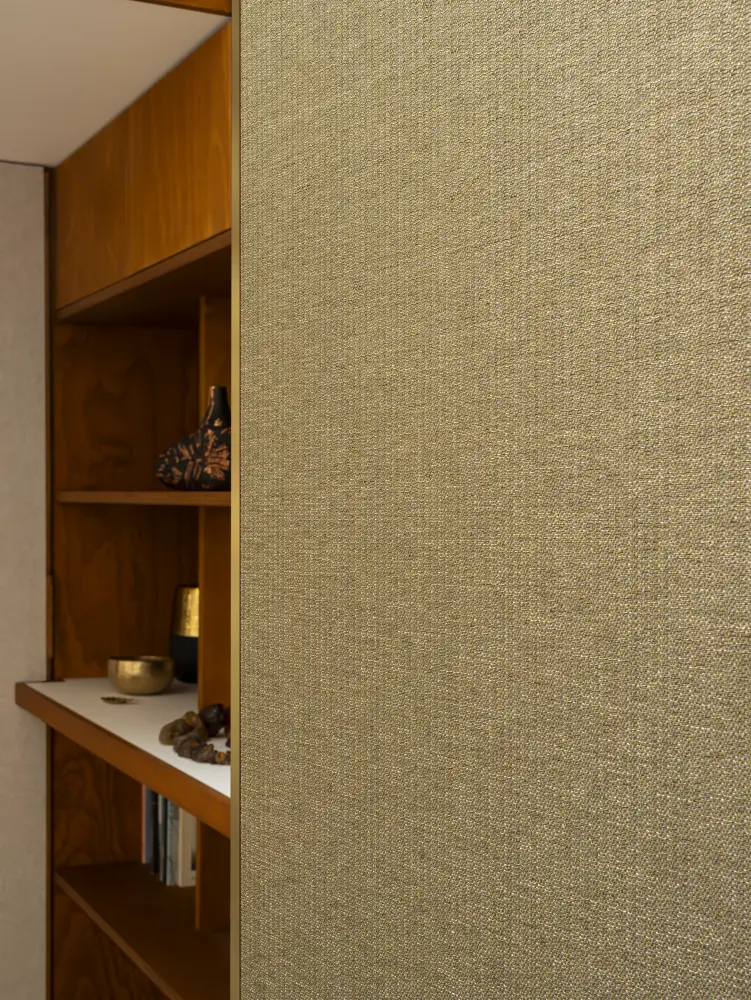 Close-up of Vescom Amber woven textile wallcovering with warm gold tone and irregular melange texture beside wooden shelving