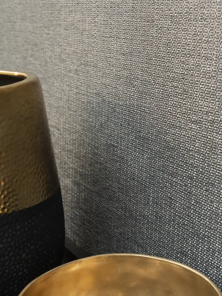 Close-up of Vescom Amber woven textile wallcovering with pronounced weave and irregular melange beside a brass bowl and dark vase.