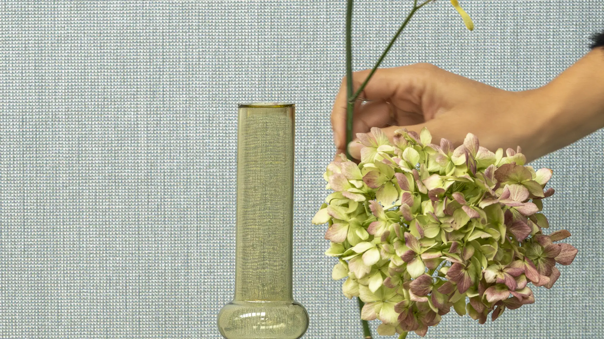 Vescom Gemstone woven textile wallcovering with small grid texture behind glass vases while a hand arranges flowers